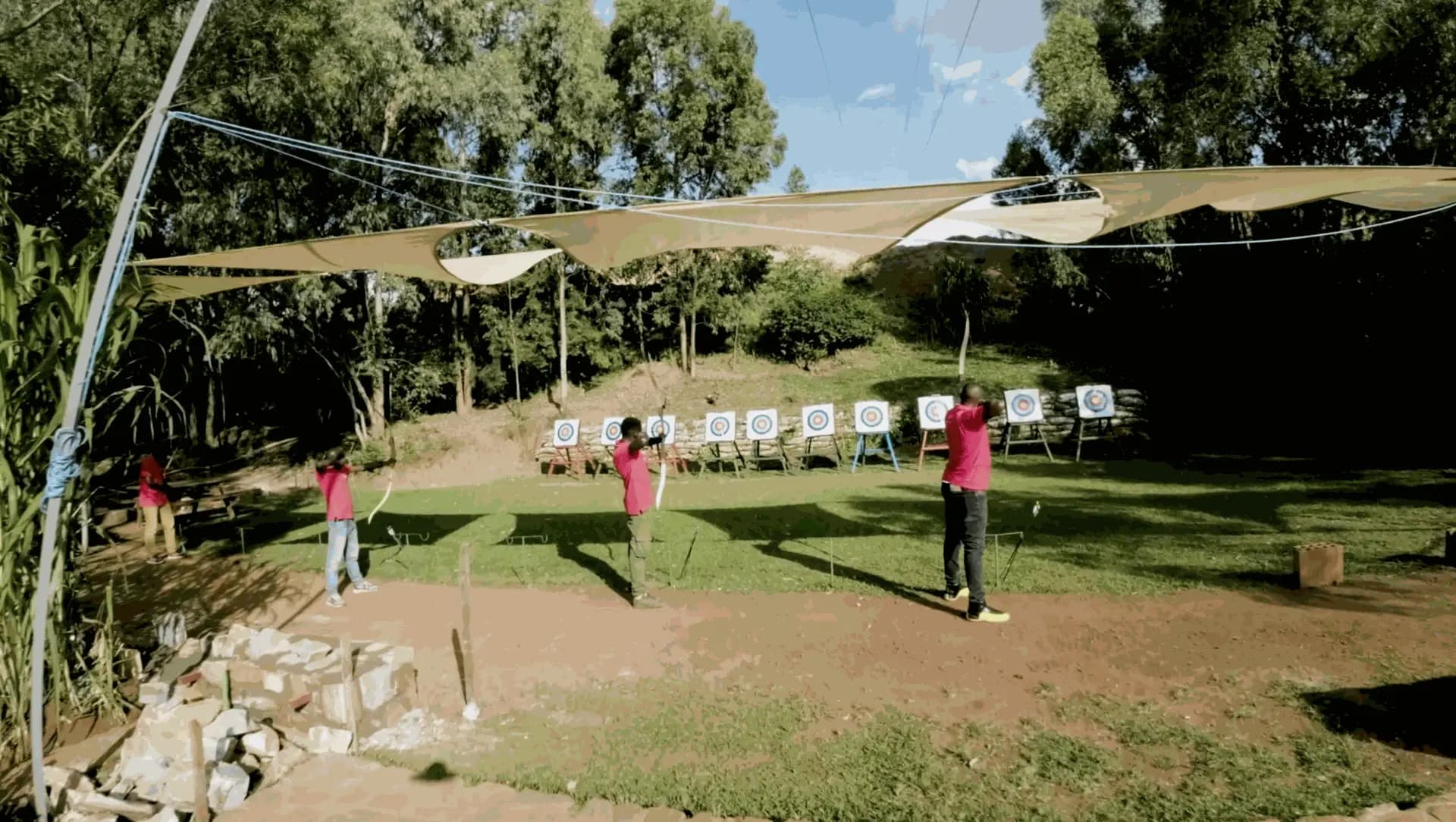 Three visitors in red shirts shooting at a line of targets on the shaded archery range.
