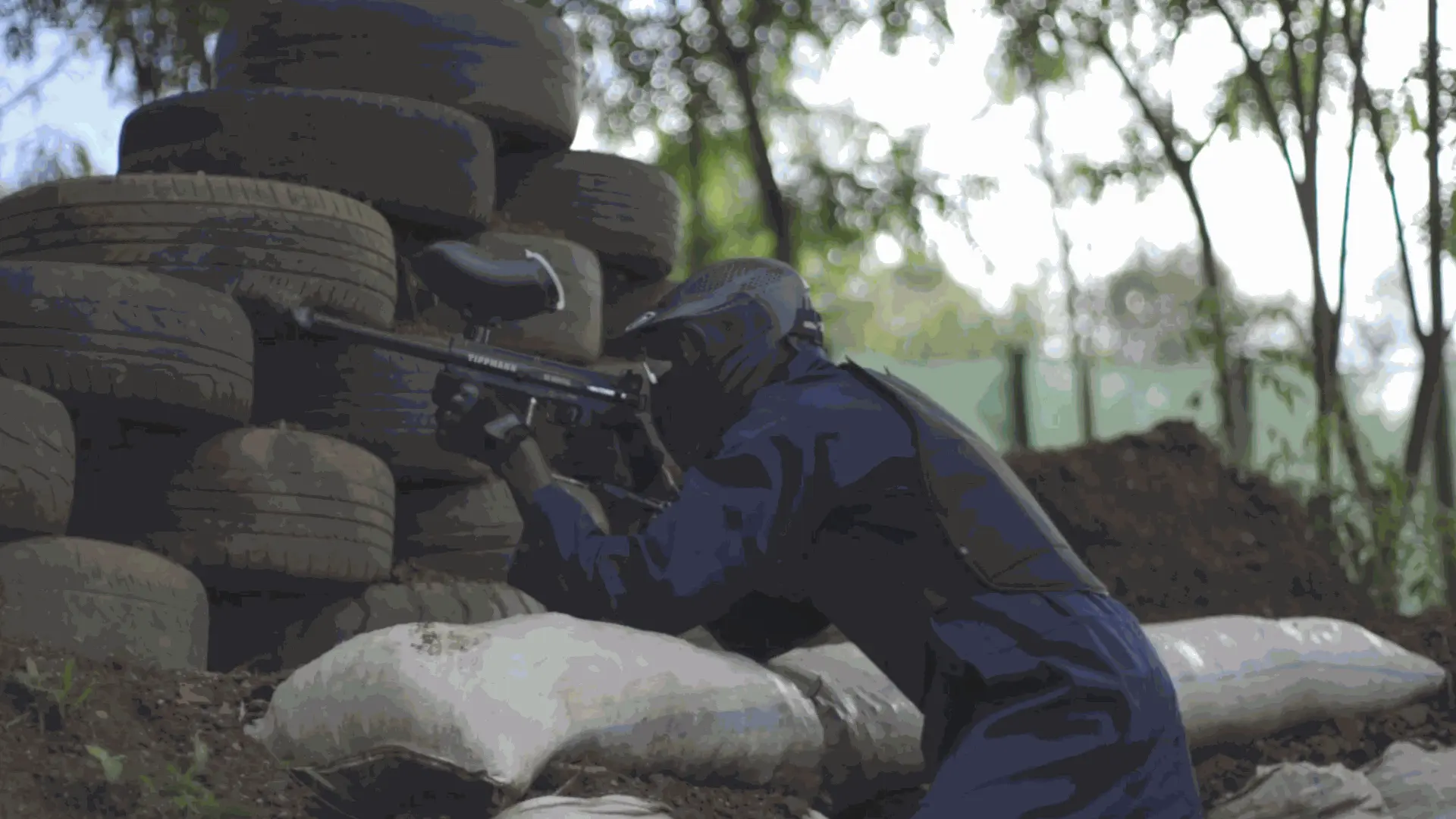 A masked paintball player aiming a marker over a tire barricade during a team battle.