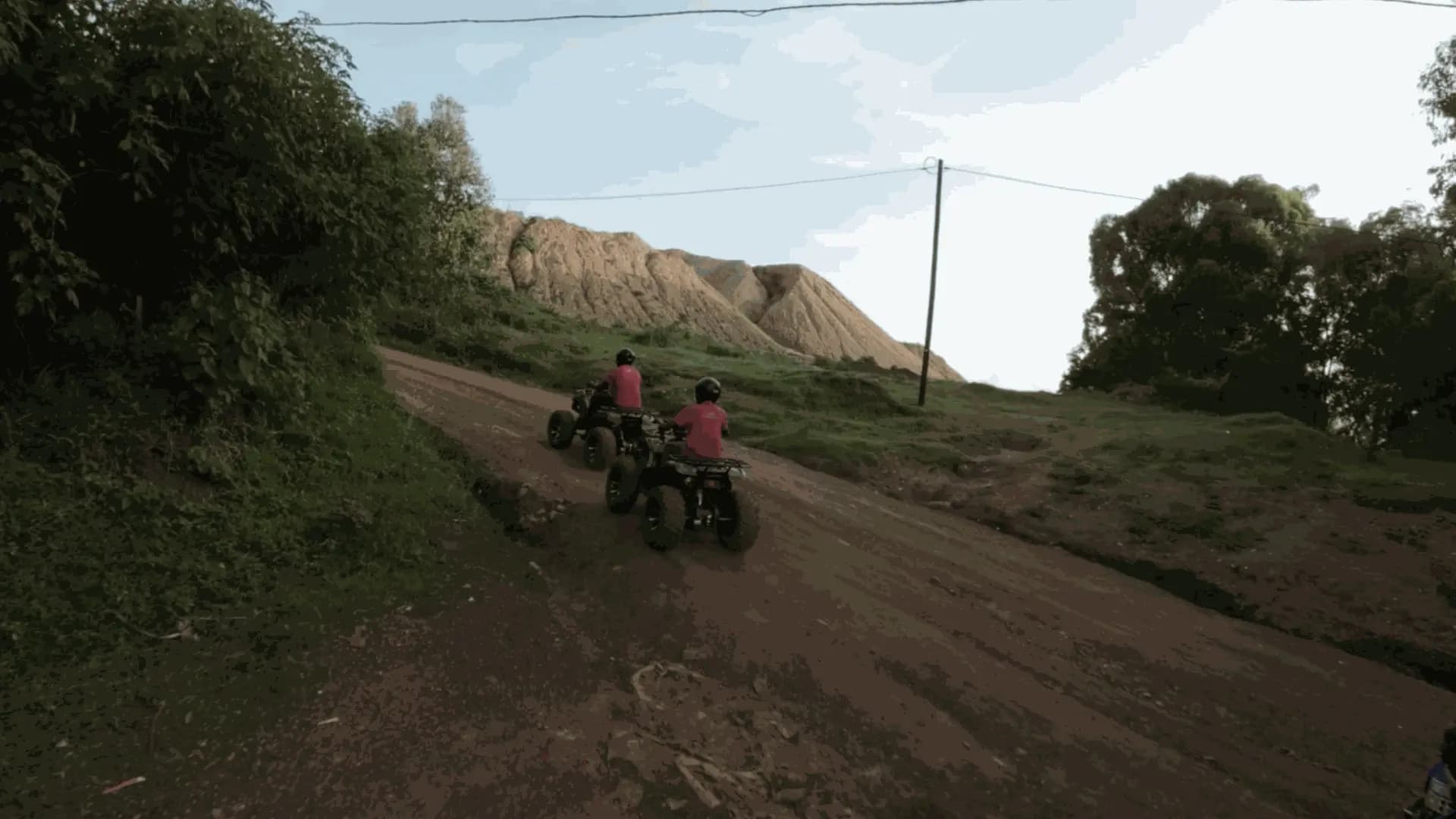 Two riders on quad bikes heading up a red dirt trail past an eroded hillside.